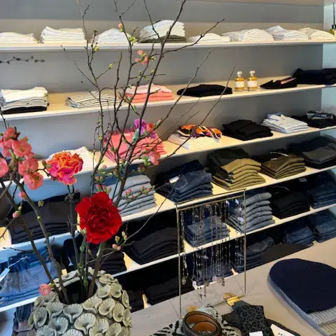 Ein Regal in einer Modeboutique mit Hosen und Oberteilen. Im Vordergrund eine Vase mit Blumen und Strauch.
